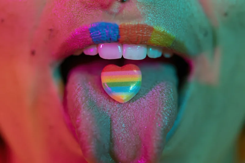 Woman with open mouth showing tongue holding rainbow heart candy, face painted in neon colors