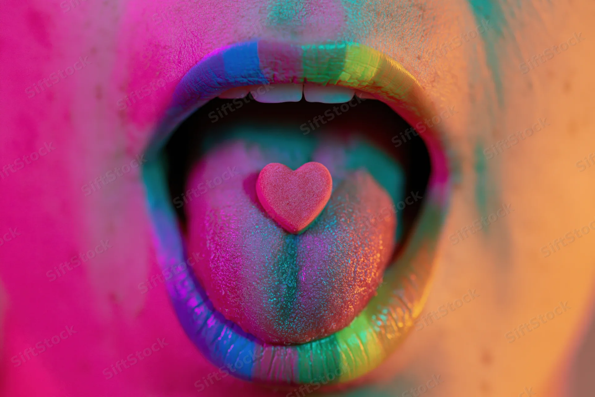 Close-up of a woman's open mouth with tongue extended showing a small heart-shaped candy on the tongue, surrounded by colorful glitter makeup