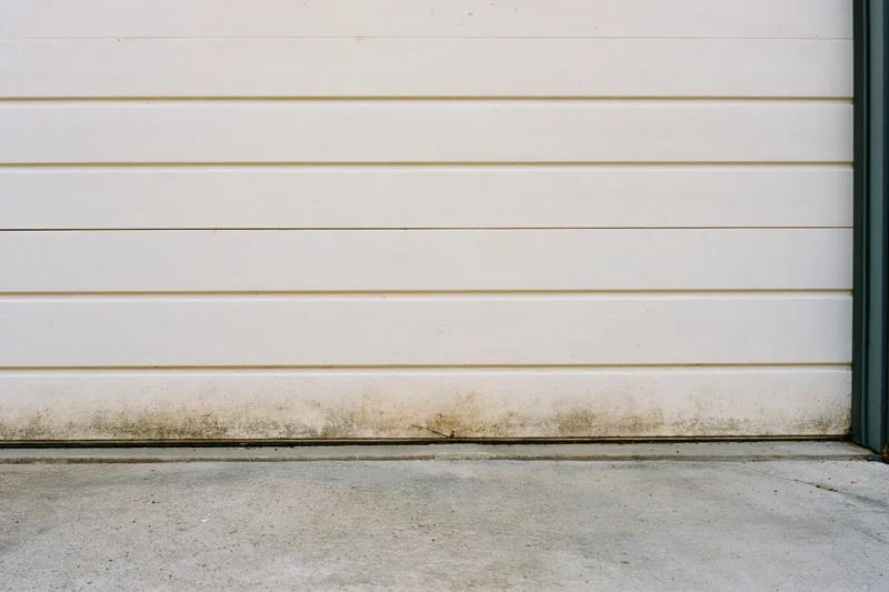 Close-up of an old off-white sectional garage door with horizontal panels and stained base against a concrete foundation