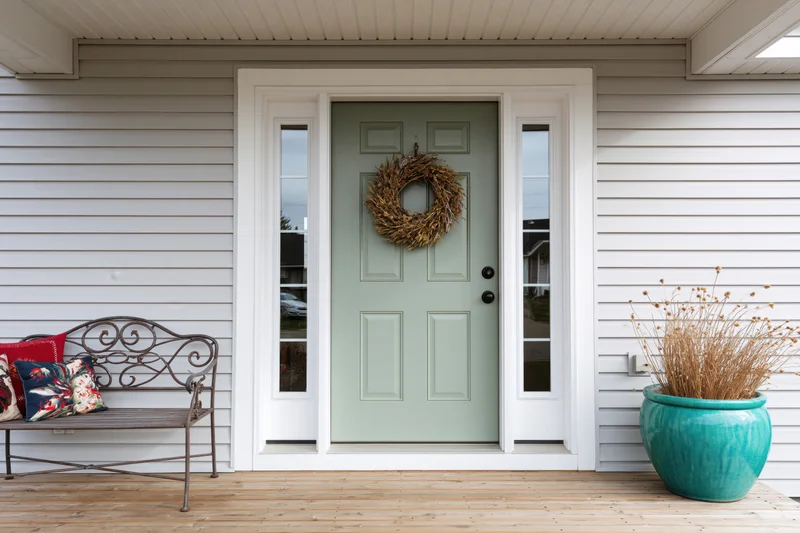 Sage green front door with dried wreath on suburban home porch, flanked by metal bench with floral pillows and turquoise ceramic planter with dried plants