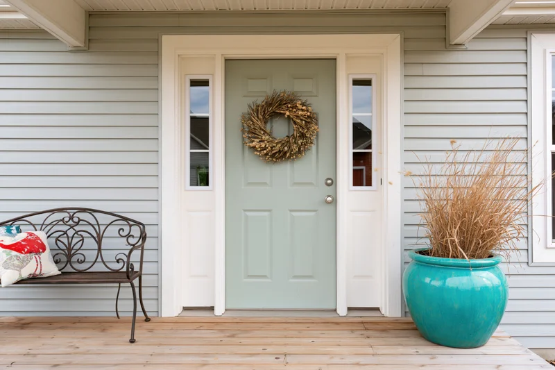 Sage green front door with dried wreath and turquoise ceramic pot on wooden porch beside metal bench with cushion