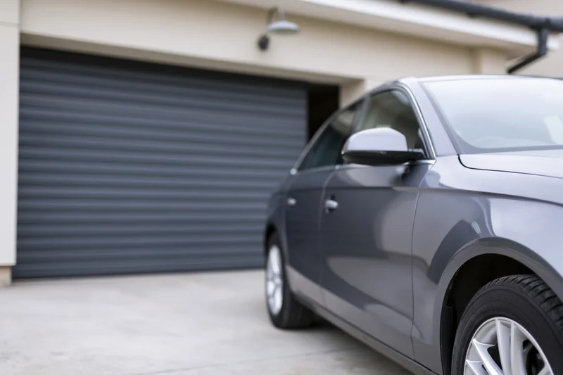 Dark grey sedan parked in front of a residential garage with closed roller door on concrete driveway
