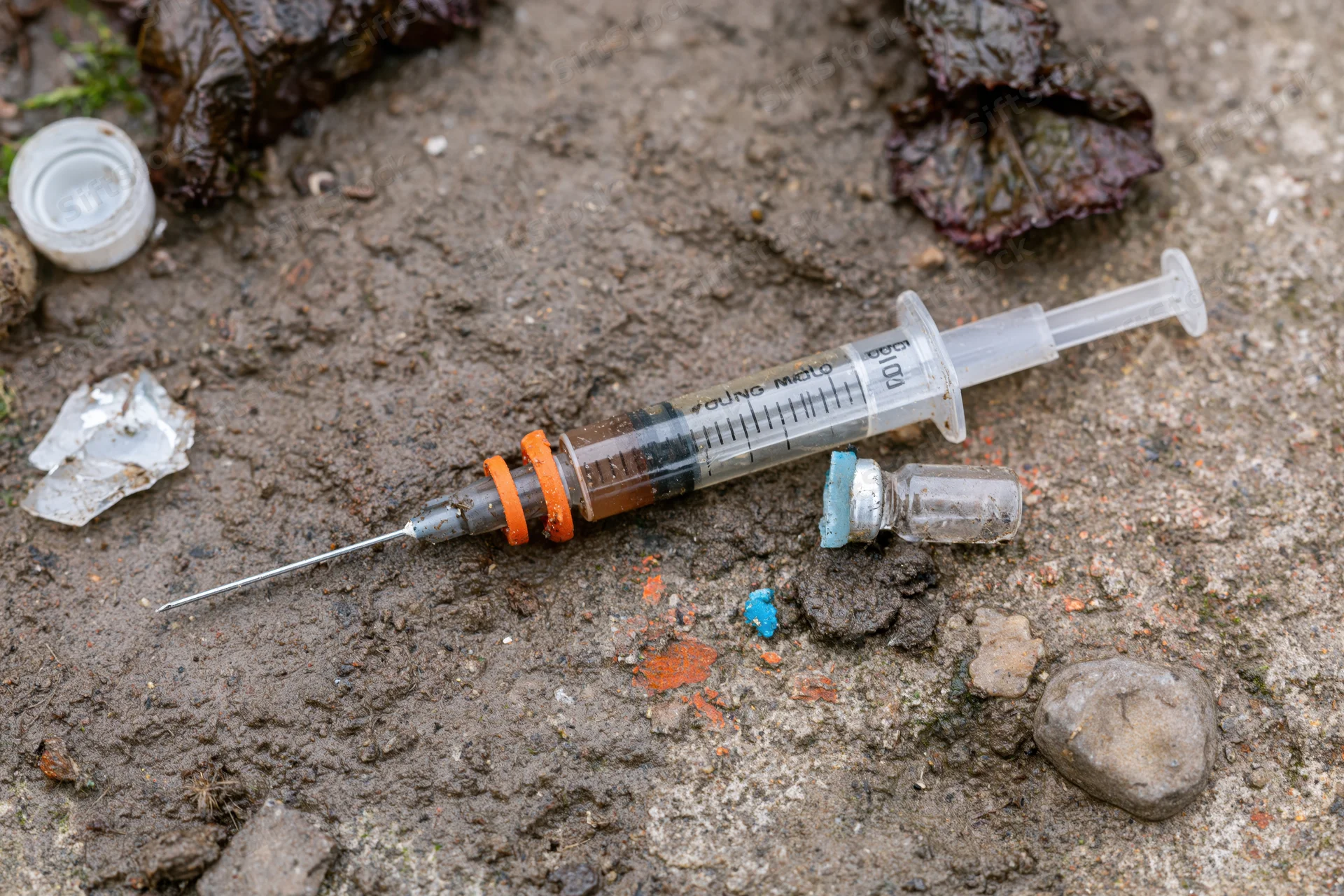 Used syringe with orange cap ring and small glass vial lying on muddy ground among debris