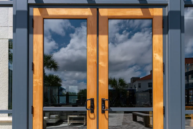 Double glass doors with wooden frames reflecting sky and clouds outside a modern building entrance
