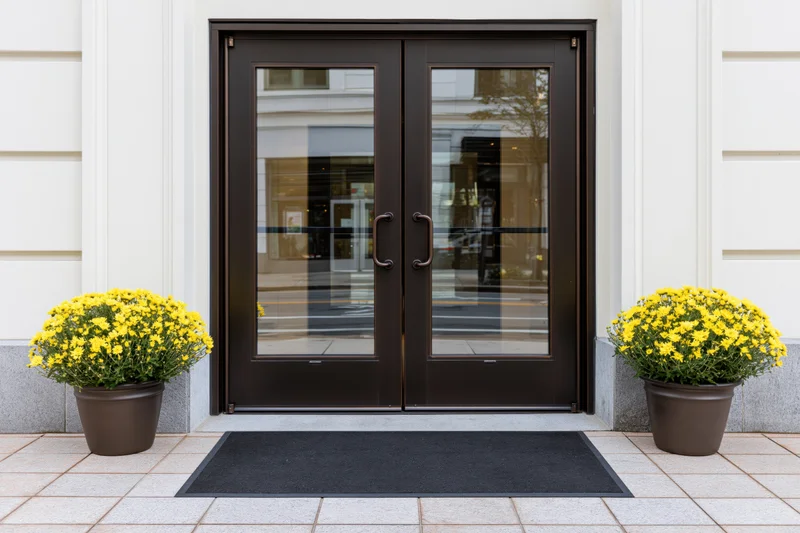 Double glass entrance doors with dark bronze frames flanked by yellow chrysanthemums in planters on a tiled sidewalk