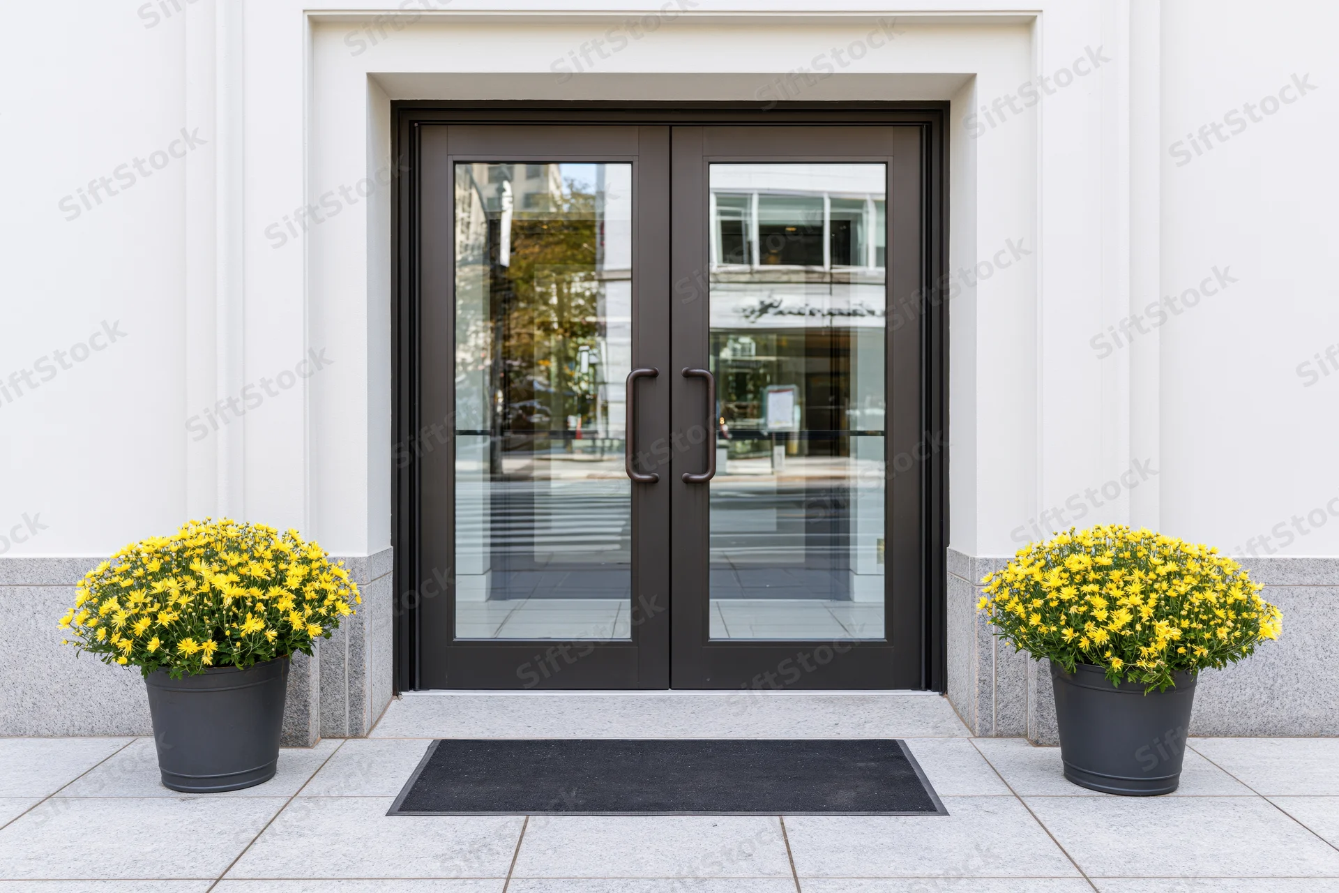 Double glass entrance doors with dark bronze frames flanked by yellow chrysanthemums in planters on a tiled sidewalk
