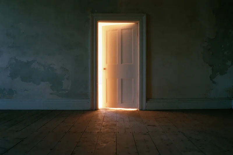 Ajar white door emitting warm golden light in a dark empty room with peeling walls and wooden floor