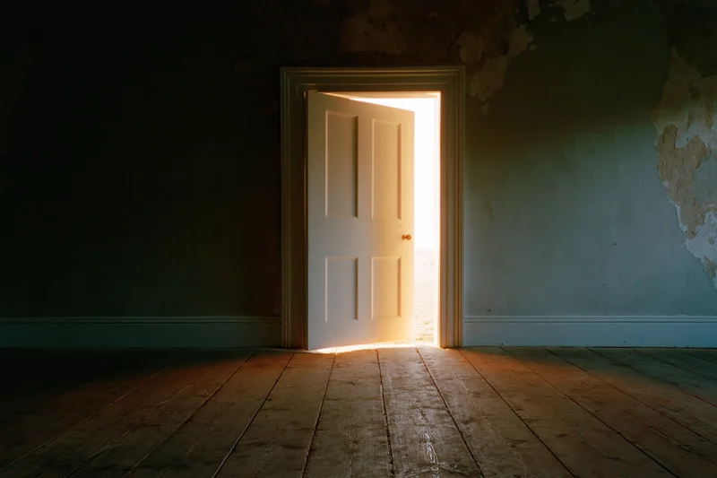 Ajar white door letting in golden light into dark empty room with peeling walls and wooden floor