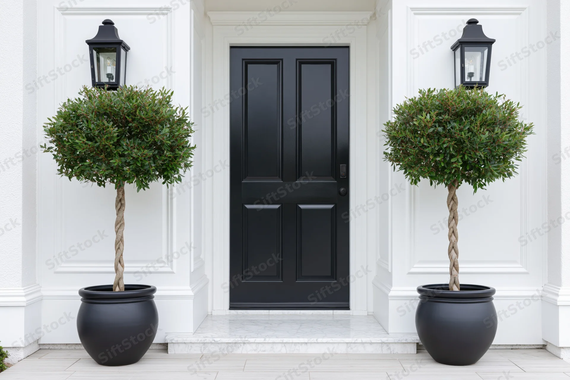 Black front door of an upscale home flanked by potted topiary trees and wall lanterns on a white facade