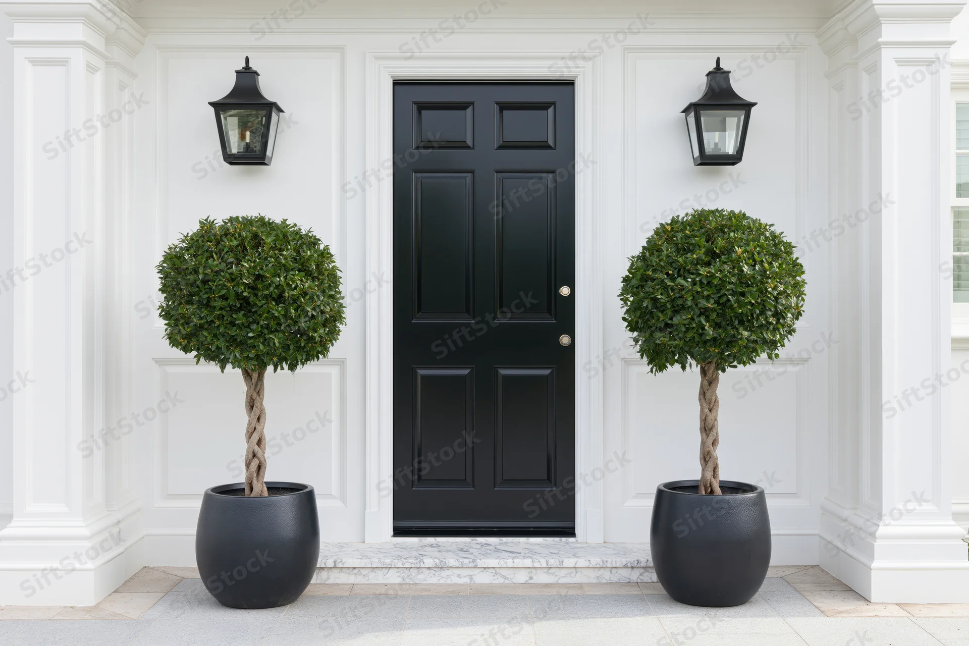 Black front door of an upscale residential home flanked by symmetrical topiary plants in black pots under wall lanterns