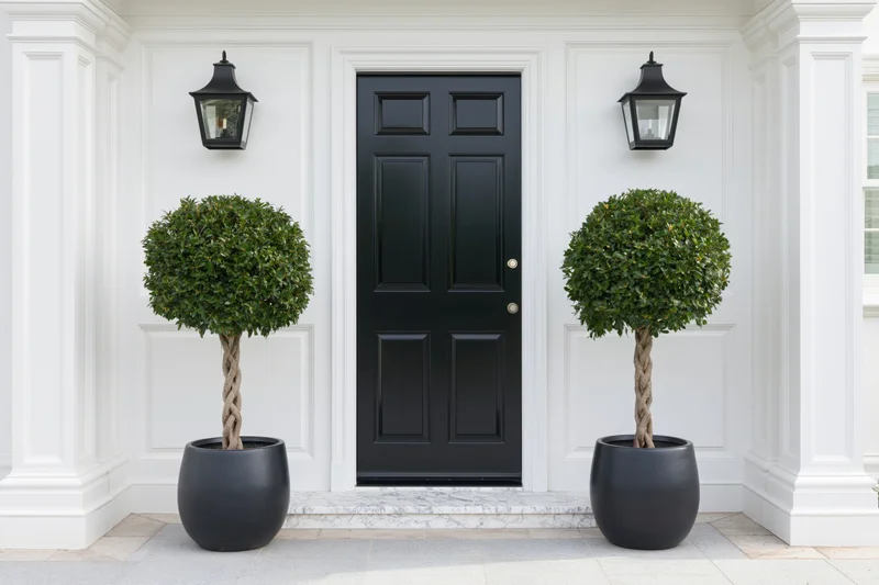 Black front door of an upscale residential home flanked by symmetrical topiary plants in black pots under wall lanterns