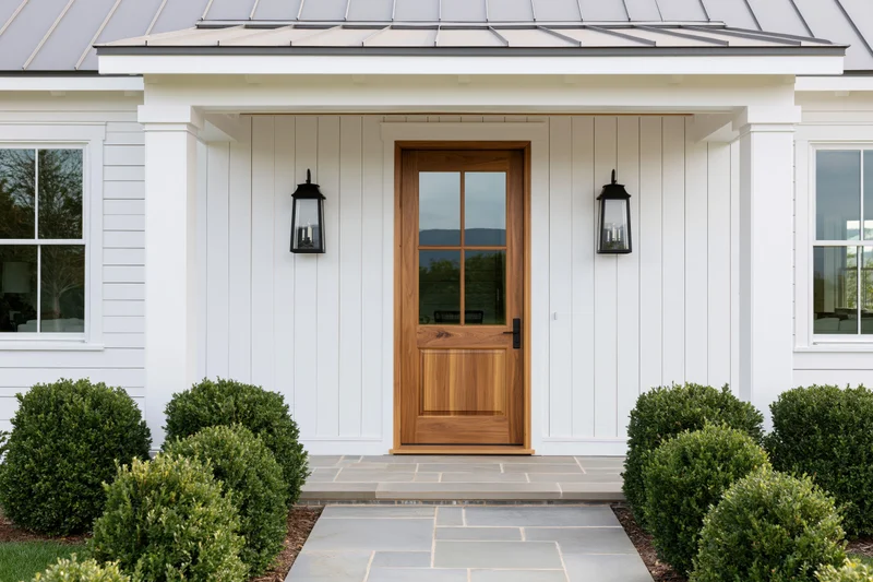 Modern farmhouse front entrance with wooden door and white siding flanked by lanterns and shrubs