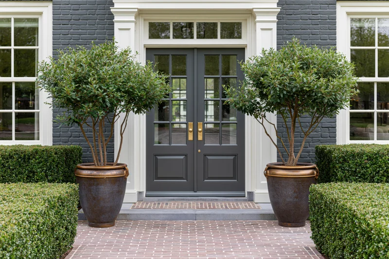 Double gray front door with copper planters and topiary trees on a brick pathway flanked by hedges