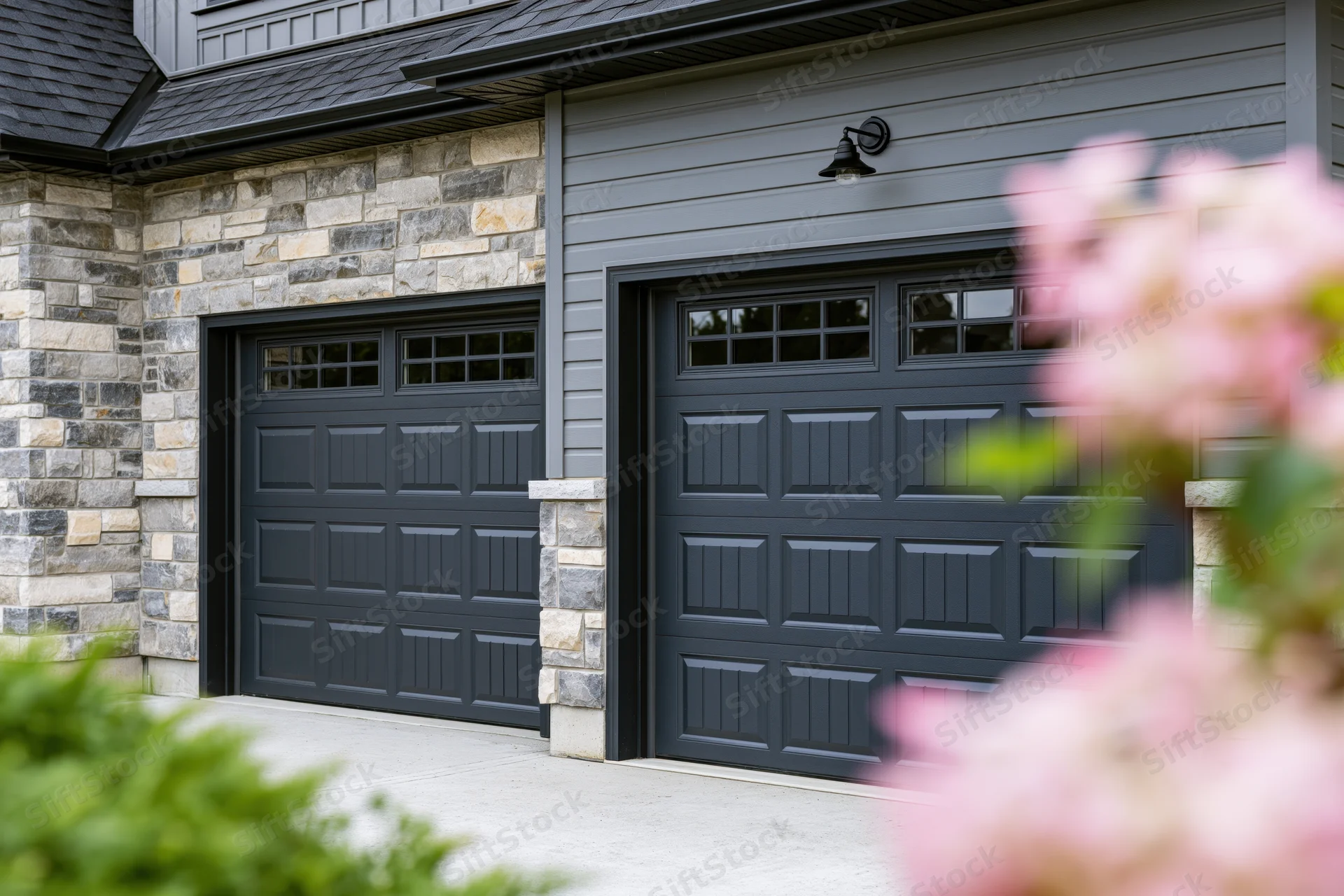Two dark charcoal garage doors on a residential home exterior with stone and siding, framed by pink flowers and greenery