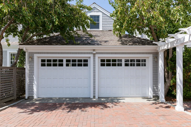 Two-car garage with white raised-panel doors and brick driveway in front of a suburban house