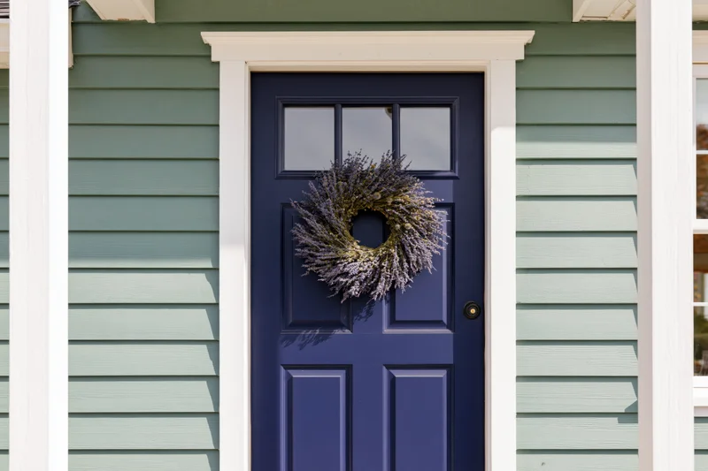Navy blue front door with lavender wreath on green siding house exterior
