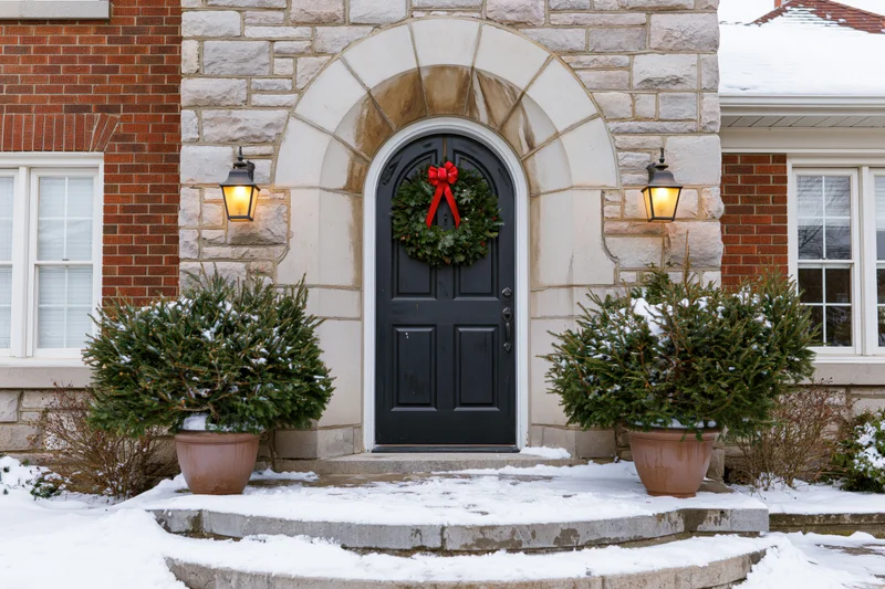 Front entrance of a stone and brick house with a christmas wreath on the door, flanked by potted evergreens in snow