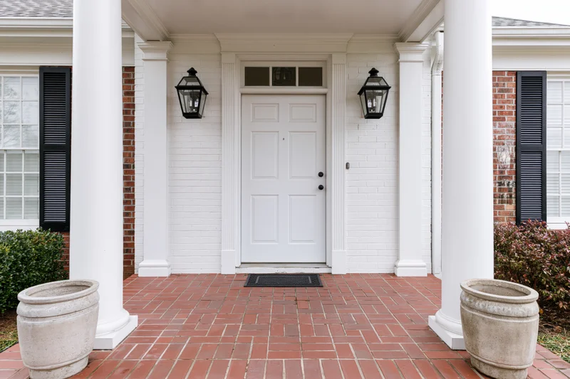 White paneled front door centered under a portico with white columns and flanked by black lanterns on a brick porch with planters