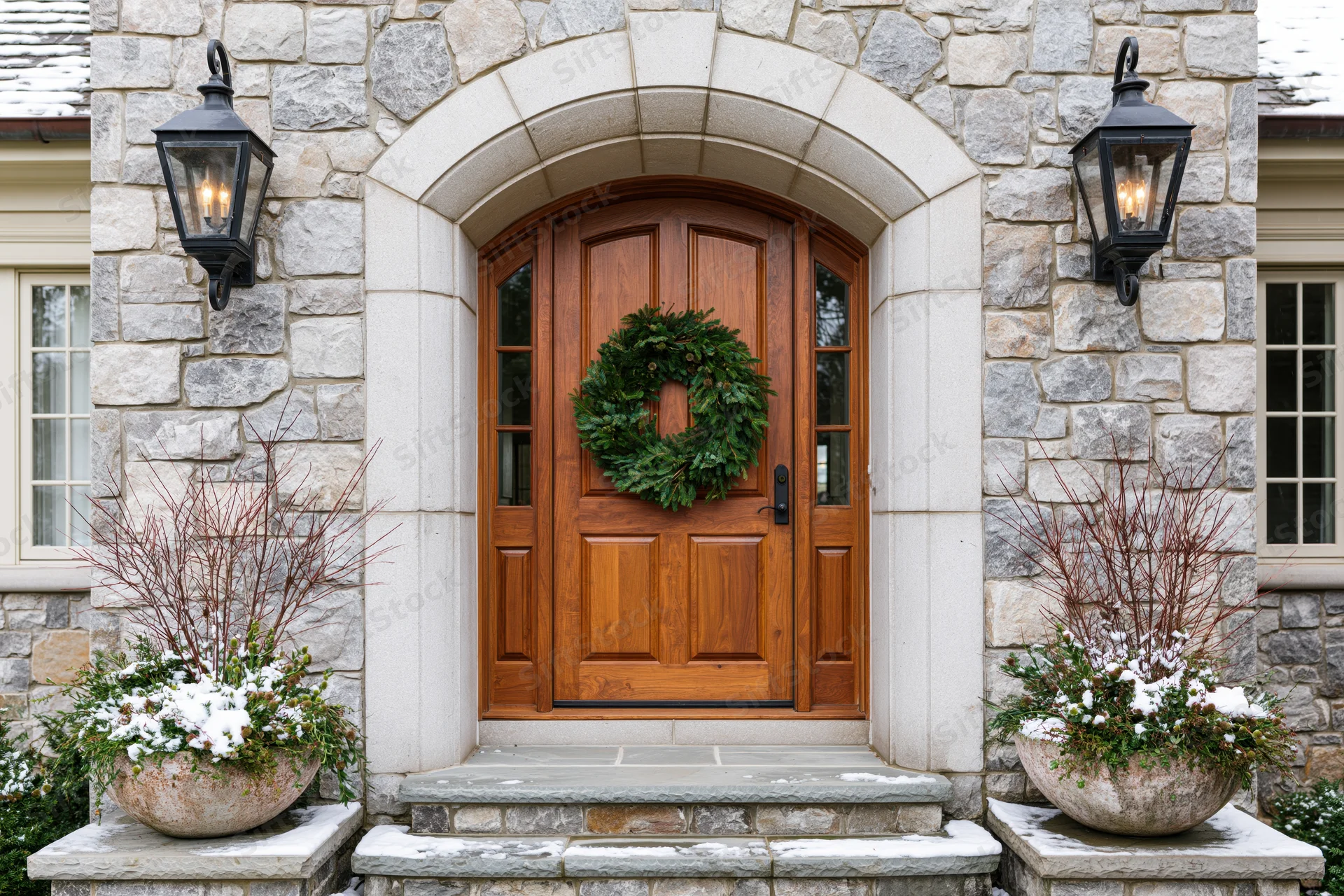 Front entrance of a stone house with a wooden door, christmas wreath, and snow-covered planters flanked by lanterns