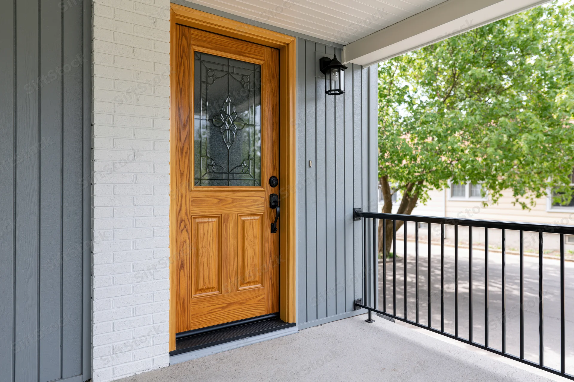 Wooden front door with stained glass panel on a suburban home porch under overhang with gray siding and black railing