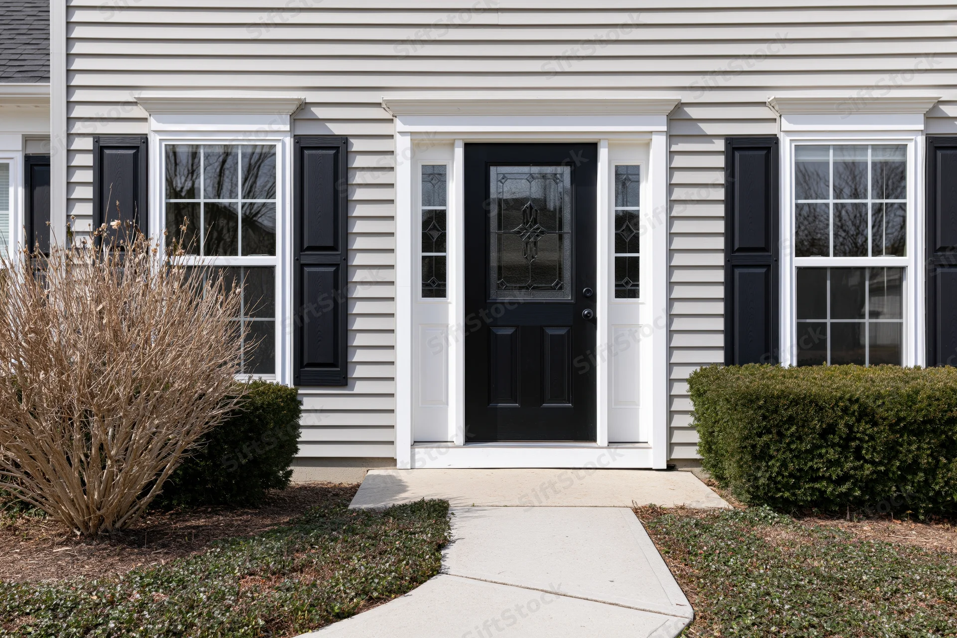 Front exterior of a suburban american colonial-style house with black door and white trim, flanked by windows and shrubs, leading to a concrete pathway