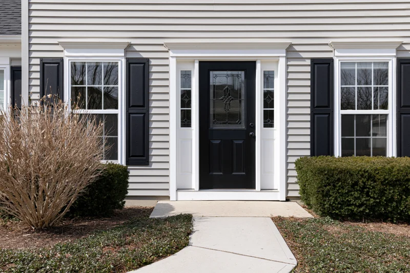 Front exterior of a suburban american colonial-style house with black door and white trim, flanked by windows and shrubs, leading to a concrete pathway