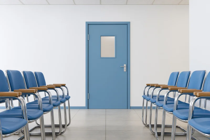 Rows of blue chairs facing a pale blue door in a hospital waiting room corridor