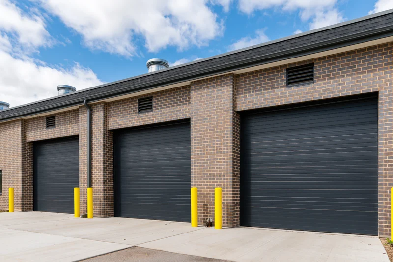 Modern brick commercial building with three dark garage doors and yellow bollards under a blue sky