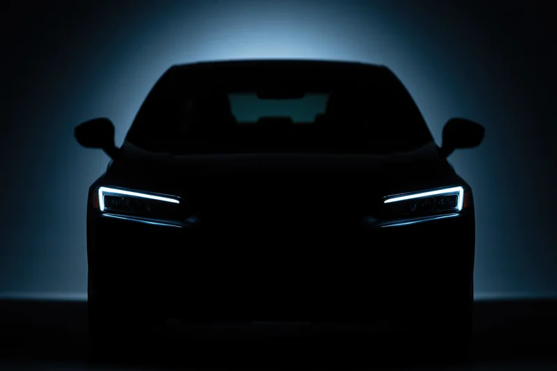 Modern sedan silhouetted head-on in near-total darkness with illuminated headlights