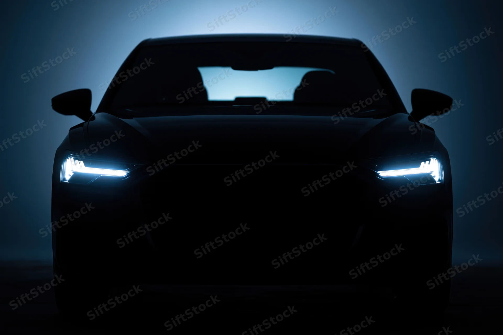 Modern sedan silhouetted head-on in dark studio with glowing led headlights