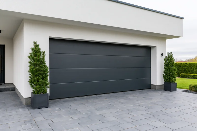 Modern single-story house exterior with dark charcoal garage door and gray stone driveway flanked by evergreen trees in planters