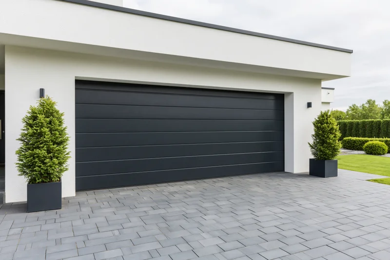 Modern single-story house exterior with large dark charcoal garage door and paved driveway flanked by shrubs in planters