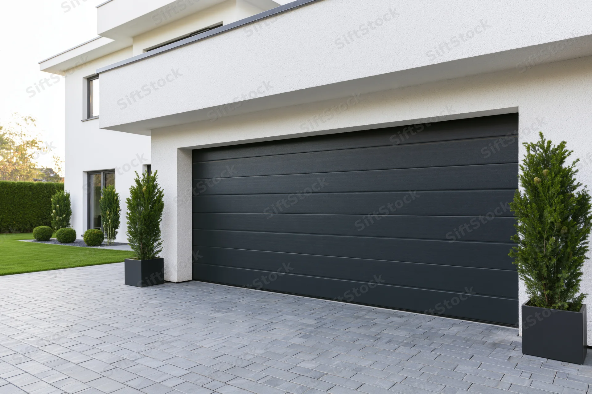 Modern single-story house exterior with large dark charcoal garage door and paved driveway flanked by potted evergreen plants