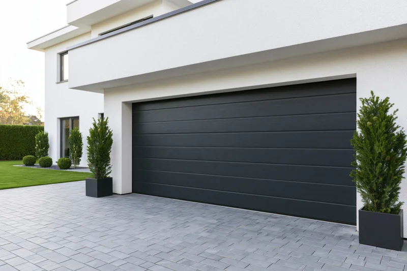 Modern single-story house exterior with large dark charcoal garage door and paved driveway flanked by potted evergreen plants