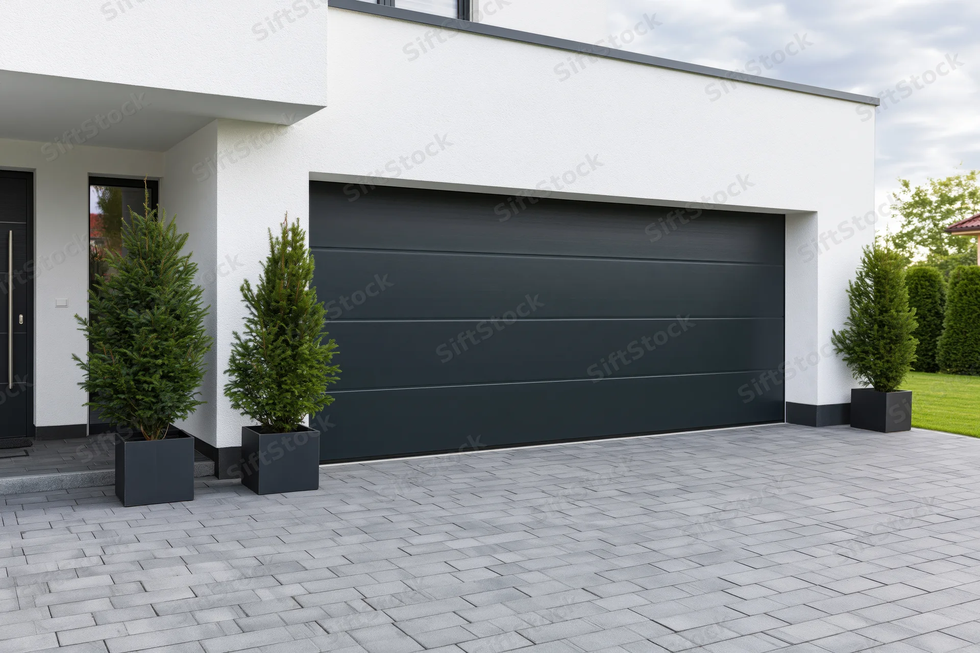 Modern single-story house exterior with dark charcoal garage door and potted evergreens on paved driveway