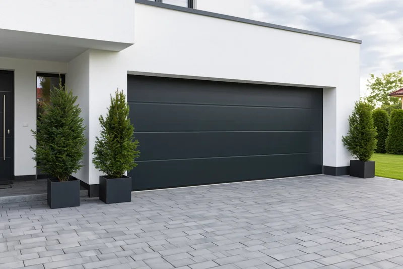 Modern single-story house exterior with dark charcoal garage door and potted evergreens on paved driveway