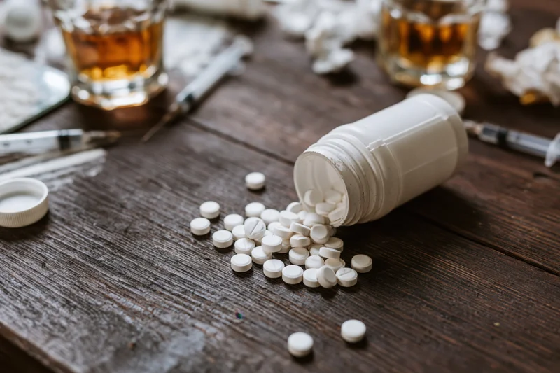 Overturned pill bottle spilling white tablets onto a dark wooden table with syringes and glasses of whiskey nearby