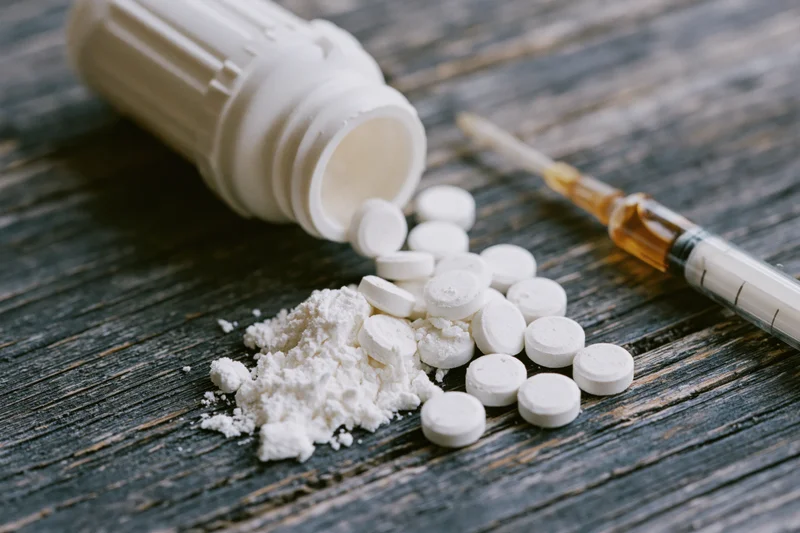 Scattered white pills and powder from an overturned bottle with a syringe on a wooden surface