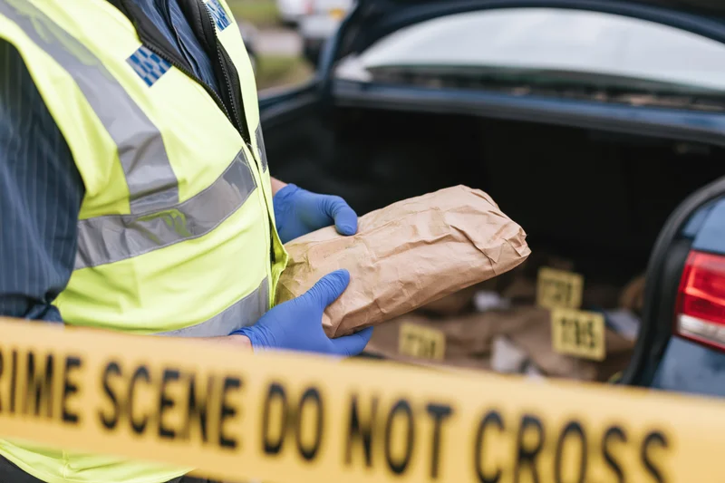 Police officer holding evidence bag near crime scene tape by open car trunk