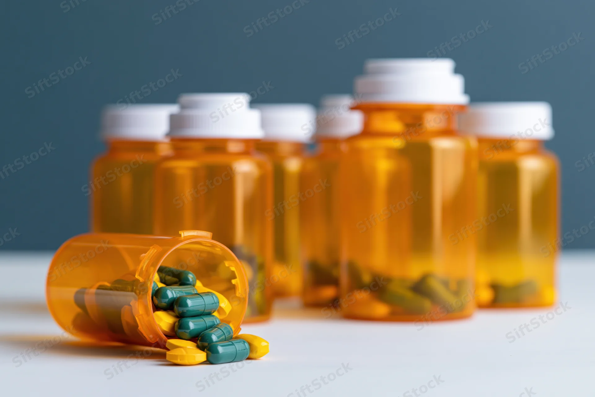 Several orange prescription pill bottles arranged on a white surface with one tipped over spilling green and yellow capsules