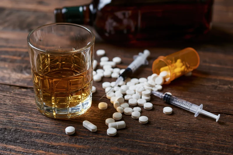 Whiskey glass and scattered prescription pills with syringes on wooden table