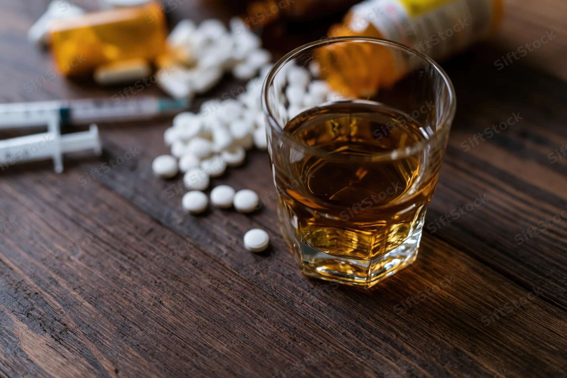 Whiskey glass with scattered prescription pills and syringe on wooden table
