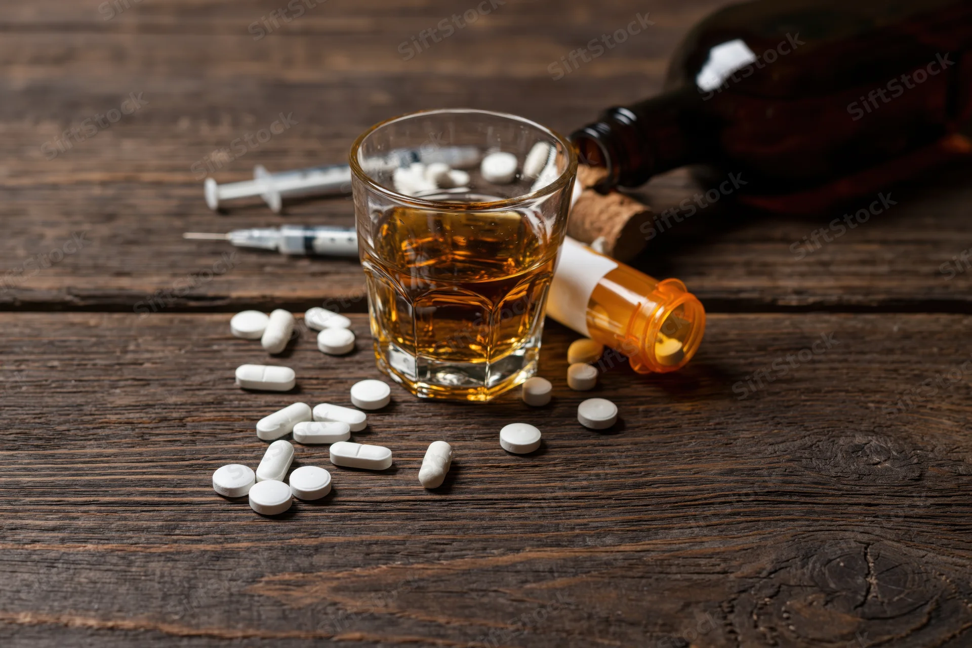 Whiskey glass with scattered prescription pills and syringes on a dark wooden table
