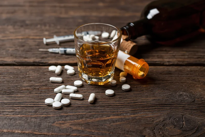 Whiskey glass with scattered prescription pills and syringes on a dark wooden table