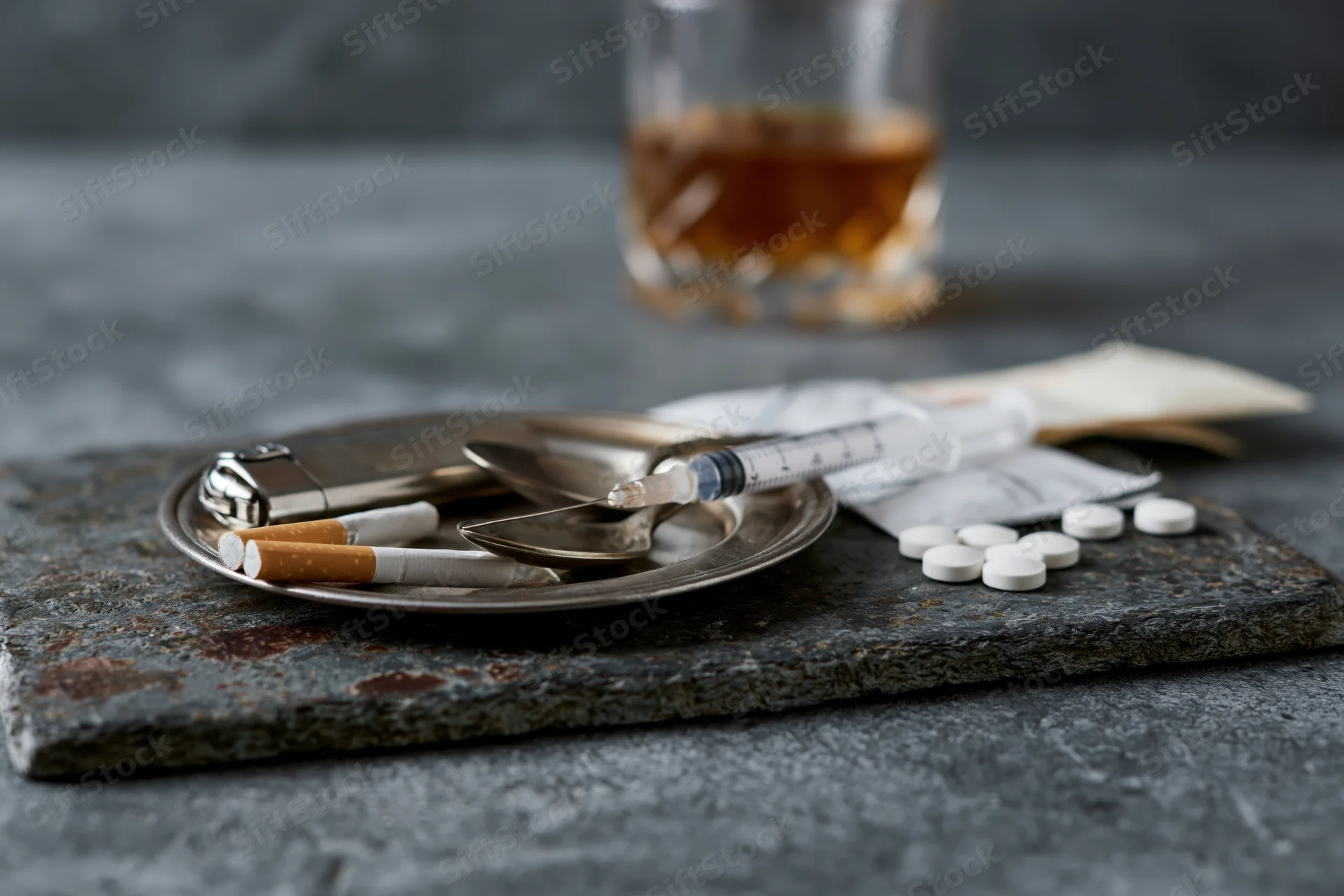 Substance abuse paraphernalia arranged on a dark slate tray with syringe, pills, cigarettes, and glass of whiskey in background