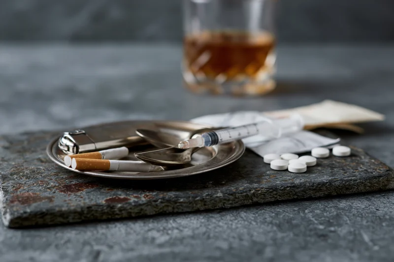 Substance abuse paraphernalia arranged on a dark slate tray with syringe, pills, cigarettes, and glass of whiskey in background