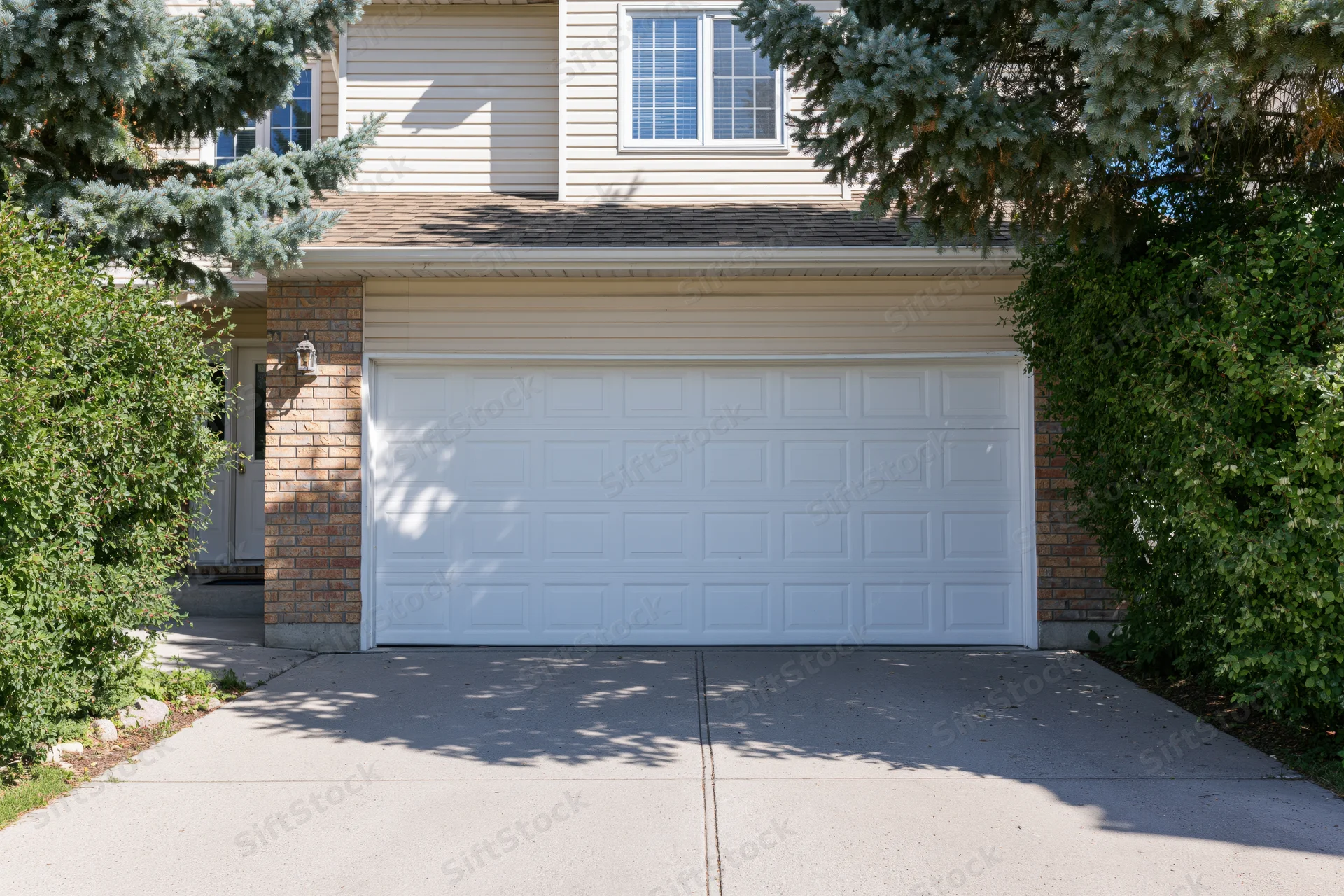 Suburban two-story house exterior with attached double garage and driveway flanked by evergreen trees and shrubs