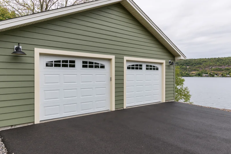 Two-car attached garage with white doors and sage siding on a craftsman-style home overlooking a lake