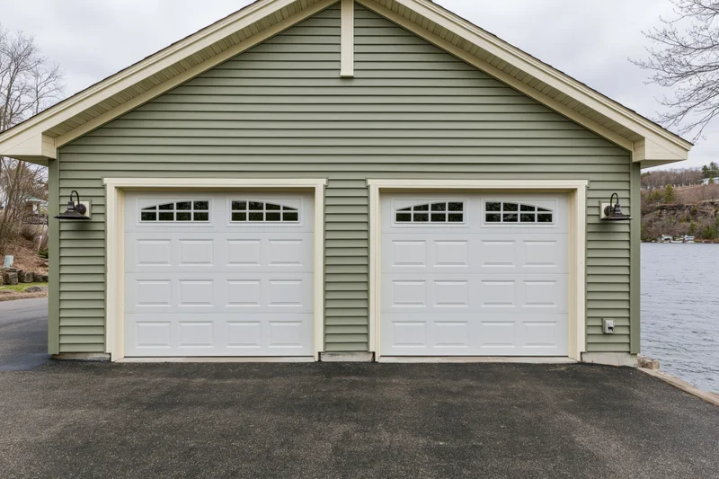 Two-car attached garage with sage siding and white doors on a craftsman-style home by a lake