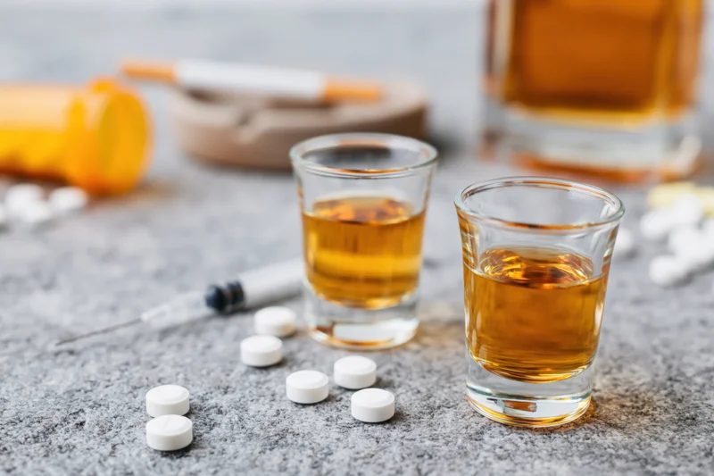 Two shot glasses of amber whiskey on a gray stone surface with pills, syringe, cigarette and ashtray scattered around